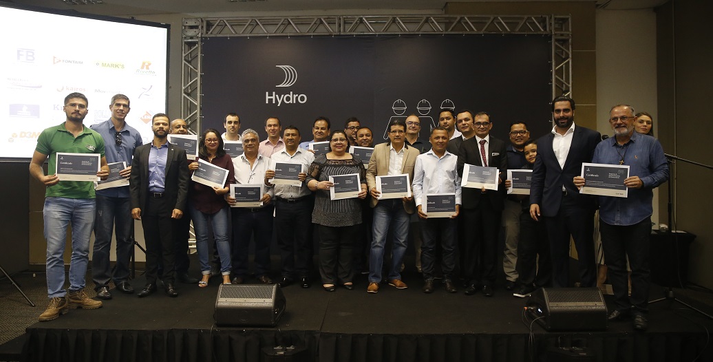 Um grupo de homens e mulheres, de pé, no palco, segurando certificados e sorrindo